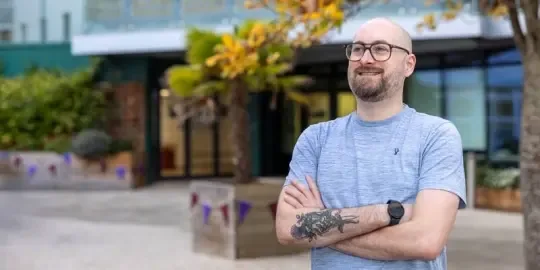 Matt, smiling with his arms folded, standing outside in front of a tree with yellow leaves.